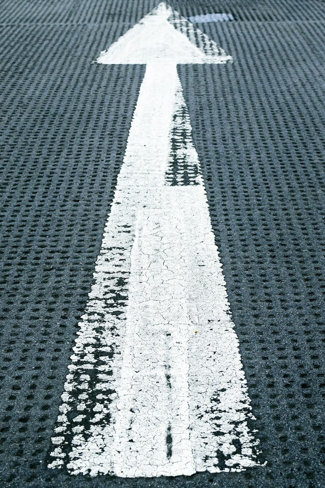 Close-up of a white directional arrow on a textured asphalt road, symbolizing forward movement and guidance.